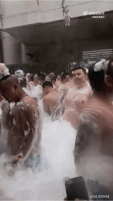 画面里一群赤裸上身的男子被大量白色泡沫包围，身上也沾满了泡沫，他们身处一个类似户外或半户外的空间，看起来像是在参与集体洗浴类活动，场景显得十分热闹滑稽，充满生活趣味。