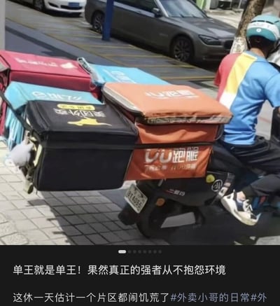 图片中一名外卖配送员正骑着电动车，车后座堆叠着多个不同平台的外卖配送箱，包括美团、饿了么、UU跑腿等，箱子数量众多几乎占据了车身后半部分。图片底部配有文字：“单王就是单王！果然真正的强者从不抱怨环境 这休一天估计一个片区都闹饥荒了 #外卖小哥的日常#外”，以幽默夸张的方式称赞该配送员接单能力强，暗示其业务量巨大。
