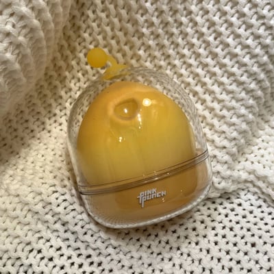 一张展示透明圆顶容器的照片，容器内装有一个黄色的圆形物体，顶部有一个黄色的小按钮。容器底部印有'PINK PUNCH'字样，整体放置在白色针织纹理的背景上。