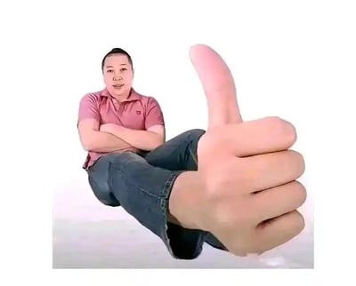 这是一张利用视觉错位打造的趣味照片：画面里的男子坐着，双臂交叉抱在胸前，画面右下角的手属于另一个人，通过精准的拍摄角度配合，让这只手和男子的腿部重合，看起来仿佛男子的腿变成了一只竖起大拇指的手，制造出荒诞又搞笑的视觉错觉，整体效果新奇有趣，没有双关、谐音梗等内容。