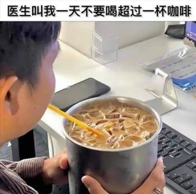 图片中一名男子侧身手持一个超大金属杯，杯中装满加了冰块的棕色冰咖啡，正用黄色吸管饮用。背景可见办公桌，上面有键盘、手机等办公物品。顶部文字写着“医生叫我一天不要喝超过一杯咖啡”，通过夸张的“一杯”容量（超大杯子）制造幽默效果，调侃人们对健康建议的“灵活”解读。