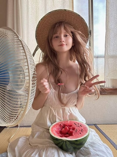 画面呈现出充满夏日氛围感的居家场景：一位有着浅棕色长卷发、戴着宽檐草帽的少女，身穿白色吊带裙，坐在铺有榻榻米的房间里。身旁的风扇正吹得她发丝飞扬，她一只手拿着盛有西瓜的勺子，腿上放着半个被挖过的西瓜，另一只手比出V字手势，脸上带着温柔惬意的笑容，整体氛围治愈又清凉，完美诠释了夏日居家的慵懒舒适感。
