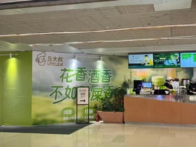 图片展示了丘大叔（UNCLE Q）奶茶店的店内场景，背景墙上方有品牌logo，中间是大幅标语，原应是宣传茶香的内容却被修改为“花香酒香不如屎香”的恶搞文字，形成强烈反差。柜台后有工作人员正在操作，上方电子屏显示饮品菜单，整体环境为商场或机场内的餐饮区，地面整洁，光线明亮。该图片通过篡改店铺招牌文字制造幽默效果，属于典型的商业场景恶作剧改编。