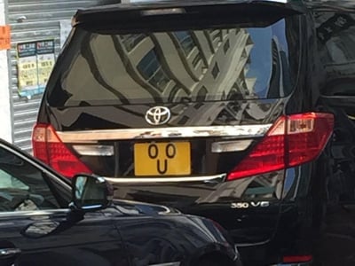 这张图片展示了一辆黑色丰田350 V6车型的尾部，其黄色车牌上的字符“O O U”巧合组成了一个类似笑脸的图案（两个“O”像眼睛，“U”像微笑的嘴巴），充满趣味性。画面左下角露出另一辆黑色轿车的侧后视镜与部分车身，背景中车辆反光能看到建筑物轮廓，还有带张贴物的卷帘门，场景为户外停车区域。