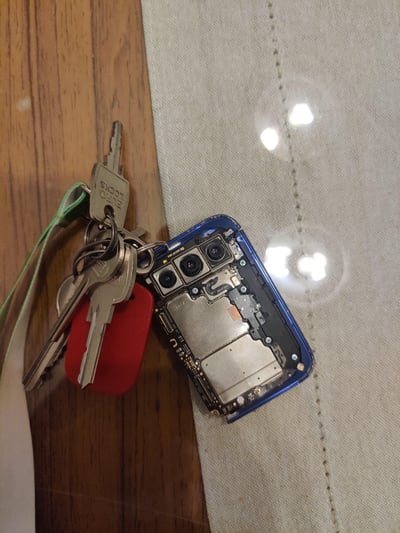 一张展示创意DIY的照片：一部蓝色智能手机的后盖被完全拆除，露出内部的三个摄像头模组和金属主板，机身被改装成钥匙扣，与一串钥匙（包含银色钥匙、红色标签和绿色挂绳）串联在一起。物品放置在木质桌面和浅色布料的交界处，光线反射在手机金属部件和布料表面，形成明暗对比。