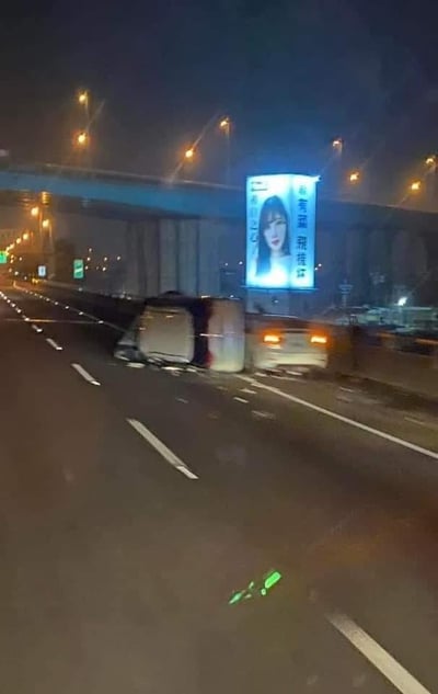 这是一张夜晚城市高架道路的实拍照片，画面核心是发生在行车道上的交通事故：一辆厢式货车侧翻横卧在路面，旁边的白色轿车停在护栏附近，路面散落着绿色的物品，现场显示出事故的突发与混乱。背景中，高架桥墩、明亮的路灯构建出夜晚城市道路的环境，一块大型亮灯广告牌格外醒目，广告牌上印有一位女性模特的人像，搭配文字“尚有菜我惜体”“有自己之心”，广告牌的文字与现场事故场景形成了巧合的视觉关联，容易引发观者的联想。