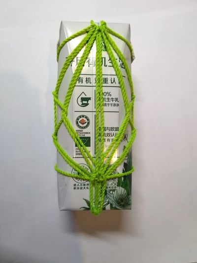 一张展示有机牛奶盒的照片，牛奶盒被鲜艳的绿色绳子以复杂的编织方式完全包裹，形成类似网兜的装饰效果。牛奶盒本身为白色基底，带有绿色有机食品相关文字和认证标志，包括'有机双重认证'、'100%有机生牛乳'以及中国与欧盟有机认证标识。整体构图简洁，背景为纯白色，突出展示了手工编织装饰与商业包装的创意结合。