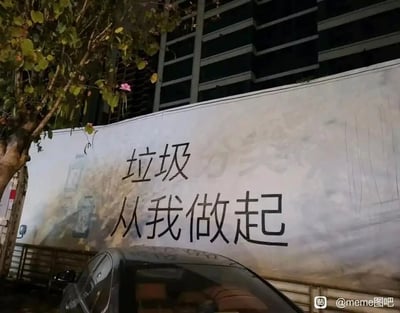 这是一张户外实拍的场景图，画面主体是一个大型宣传围挡，原本的“垃圾分类 从我做起”正向宣传标语中，“分类”二字被去除或覆盖，仅留下“垃圾 从我做起”的字样，形成了与原宣传主旨完全相反的搞笑反差效果。围挡左侧有一棵带有粉色小花的树木，下方露出一辆黑色汽车的车顶部分，背景是一栋多层建筑物，围挡右下角带有“@meme图吧”的水印标识。