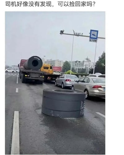 这是一张拍摄于城市雨天道路的搞笑梗图，画面中一辆载有大型钢卷的卡车行驶在前方车道，同款大钢卷掉落在后方的路面上，周围还有正常行驶的社会车辆。图片上方配文「司机好像没有发现，可以捡回家吗？」，通过调侃的语气制造反差笑点：钢卷体积庞大且重量极重，个人根本无法搬运，这种不符合现实的想法形成了幽默效果。