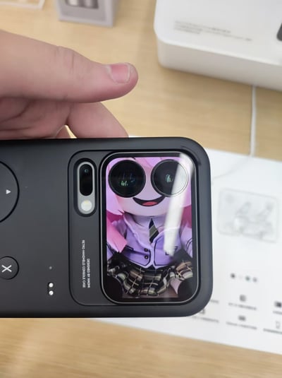 一只手握着黑色手机壳，手机壳背面的摄像头区域设计成了动漫角色的脸部，两个摄像头恰好成为角色的大眼睛。角色有着粉色头发，穿着紫色校服外套和格子短裙，面带微笑。背景是浅色木质桌面，上面还有白色的盒子和纸张。这种创意设计让手机摄像头区域变成了可爱的动漫角色脸，非常有创意和趣味性。