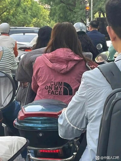 在城市拥堵的交通路口，一位女生坐在电动车后座，她的粉色外套模仿户外品牌The North Face的经典logo布局，但将品牌相关文字替换成了具有低俗含义的英文句子“CUM ON MY FACE”，形成强烈的恶搞反差感，社死效果拉满。
这是对The North Face品牌logo的恶意恶搞，利用品牌logo的视觉布局，替换成低俗英文句子，属于山寨恶搞服饰引发的社死场景。
