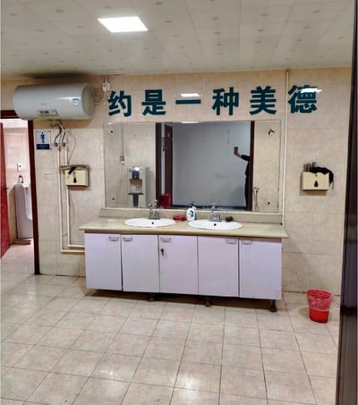 这是一张实拍的公共男卫生间洗手台区域的照片，镜子上方的瓷砖墙面上写有四个蓝色大字“约是一种美德”，原本完整的宣传标语应为“节约是一种美德”，此处缺失了开头的“节”字，意外形成了趣味玩梗效果。场景内可见双盆洗手柜、嵌在墙里的大镜子，左上角墙面安装了电热水器，左侧入口处能看到男厕标识和立式小便池，右下角地面放置了一个红色垃圾桶，镜子中反射出拍摄者举手机拍照的倒影，整体是普通的单位公共厕所装修环境。
