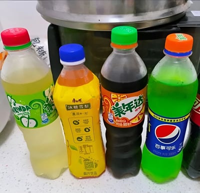 图片展示了四瓶不同品牌的饮料整齐摆放在白色台面上，背景有一个金属锅。从左至右依次为：绿色瓶盖的美年达梨味汽水（浅黄色液体，瓶身有绿色图案和“Mirinda”标识）、蓝色瓶盖的康师傅冰糖雪梨果汁饮料（橙黄色液体，瓶身黄色并印有产品名称及营养成分表）、绿色瓶盖的美年达橙味汽水（深棕色液体，橙色标签上有“美年达”字样和橙子图案）、橙色瓶盖的百事可乐（绿色液体，蓝色瓶身上有红色百事标志）。