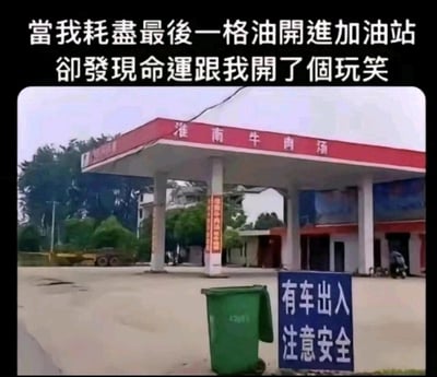 这张图片呈现了一个充满乌龙感的生活场景，顶部配文“当我耗尽最后一格油开进加油站，却发现命运跟我开了个玩笑”铺垫了搞笑的语境，画面主体是一家外观高度模仿加油站的淮南牛肉汤店铺——红白配色的顶棚、白色立柱的设计和加油站的视觉特征高度相似，会让油表见底、急于加油的司机产生误判。店铺前方立有一块蓝色的“有车出入 注意安全”警示牌，旁边还有一个绿色垃圾桶，背景能看到树木、低矮建筑和停放的电动车，整体场景是真实的户外实拍，利用建筑外观的相似性制造出强烈的反差感，精准戳中司机急于加油时的乌龙经历，充满生活趣味。