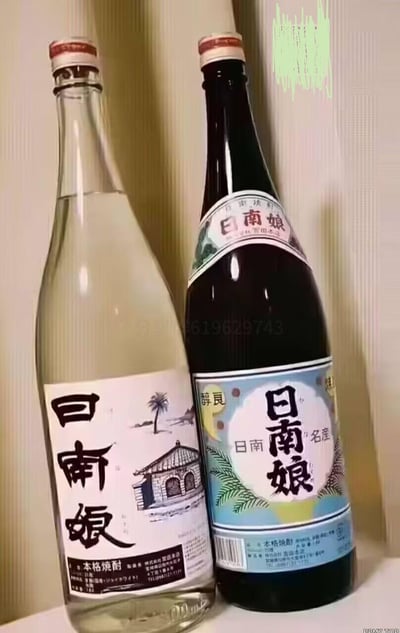 图片展示了两瓶贴有“日南娘”标签的酒类产品。左侧为透明玻璃瓶，标签印有黑色“日南娘”大字，下方配有手绘风格的建筑和棕榈树图案，底部标注“本格焼酎”等字样；右侧为深棕色玻璃瓶，蓝色底色标签上同样有“日南娘”文字及植物图案，上方可见“名産”“醇良”等字样。右上角有绿色涂鸦状痕迹，瓶身中部有模糊数字水印“619629743”，两瓶并排竖立在浅色墙面背景前。