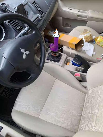 这是一张三菱汽车驾驶舱的实拍照片，原本的车辆档把被替换成了造型奇特的紫色改装件，顶部还贴了一个卡通鸭子贴纸，画风十分搞怪。车内米白色座椅搭配深色方向盘，中控与副驾驶区域堆放着纸箱、零食等杂物，整体环境略显杂乱，这个另类的档把成为了画面的绝对视觉焦点，充满趣味反差感。