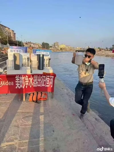 这是美的净水器的事件营销实拍场景，在以水质不佳著称的恒河岸边，品牌摆放了多款净水器产品，搭配“包干干净的”“还恒河一个‘清白’”的宣传标语，以及写着“美的净水器出征恒河”的红色横幅。一名戴墨镜的男子正在现场饮用经过净水器过滤后的恒河水，旁边有人用手机拍摄记录，通过这种极具挑战性的场景，直观展示产品的净水能力，反差感强烈，营销噱头十足。