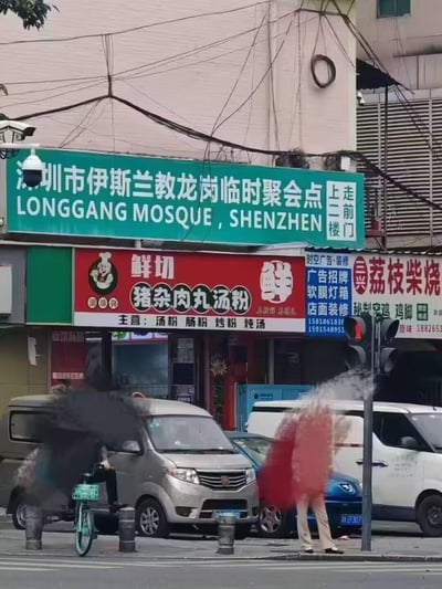 这是一张深圳龙岗街头的实拍照片，画面上方是绿色的「深圳市伊斯兰教龙岗临时聚会点（LONGGANG MOSQUE, SHENZHEN）」招牌，标注有「走前门 上二楼」的指引；下方是多家临街商铺，包括主打鲜切猪杂肉丸汤粉的餐饮店、广告装修店和荔枝柴烧熟食店。街道上有行人，两人的雨伞被风吹得严重变形，还有停放的车辆与交通信号灯，呈现出充满生活气息的日常城市街景。