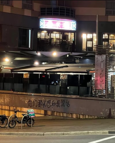这是一张夜晚的原始实拍横图，画面展现了一处城市临街商业区域的场景。上方是亮着灯光的商业建筑，带有中式风格的窗格，右侧立柱上有“DIXON NUMBER ONE”的标识；下方的矮墙表面被人用黑色字体涂写了“他们都不想和我说话”的文字，传递出落寞孤独的情绪；画面左下角停放着两辆电动车，商业场所的暖光与涂鸦的低落文字形成鲜明反差，烘托出城市里的小众情绪表达。