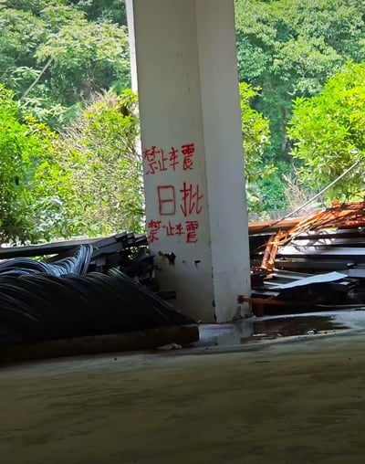 这是一张实拍的半开放建材堆放场地照片，画面中心是一根白色混凝土立柱，立柱上用红色喷漆手写了三行警示语，因书写者字体不规范、字形结构拆分错误，原本想要表达的“禁止震动、审批、禁止震动”被误写，看起来类似低俗内容“禁止车震、日批、禁止车震”，形成了荒诞的歧义效果。立柱两侧堆放着黑色波纹管材、金属板材等建材，后方是茂密的绿色阔叶树木，地面为水泥质地，整体场景为野外或城郊的建材存放区域。