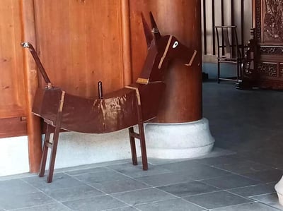 图片展示了一件放置在传统中式建筑内的木制驴形雕塑。雕塑采用深棕色木材制作，造型简约古朴，具有明显的使用痕迹和磨损。雕塑被放置在一根巨大的木柱旁，背景中可以看到传统的木制家具和雕花装饰，地面铺设着灰色石板。整个场景呈现出浓厚的中式传统文化氛围。