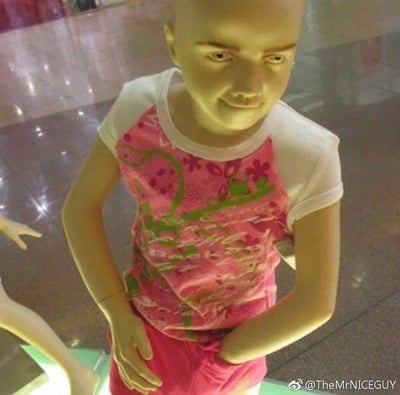 这是一张在商场橱窗拍摄的照片，画面主体是一个穿着粉色童装的儿童服装模特假人。由于视觉错位，假人的手臂与身体位置重合，看起来做出了非常怪异的姿势，搭配假人脸上诡异的微笑，整体画面充满了荒诞的搞笑感，属于偶然捕捉到的趣味视觉错位场景，没有双关或谐音梗，仅靠错位效果制造笑点。