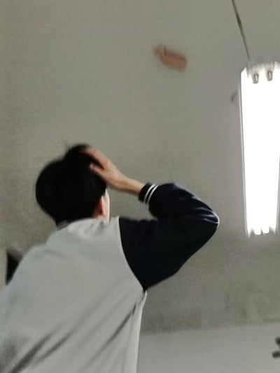 图中显示一名短发男子站在室内，背对着镜头，正抬手挠着头看向墙壁。男子穿着一件灰黑相间的夹克，房间墙壁为浅色，右上角有一扇窗户透进光线。墙壁上方有一块明显的棕色污渍，男子的动作和姿态显示出他对这个污渍可能感到困惑或烦恼。