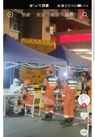 这是一张竖屏手机截图，呈现的是深夜的户外防疫核酸检测现场。画面左侧，身着全套白色防护服的医护人员正专注地为一名坐着的橙色救援服人员进行核酸采样；旁边还有三位身着同款橙色救援服的人员，衣服上印有“中国吉林消防”的字样，他们有的站立等待，有的在现场值守。现场搭建了蓝色的遮阳棚，摆放着白色桌椅，并用红白相间的隔离带划定了检测区域，背景中能看到一辆带有“急援”字样的黄色救援车辆。界面顶部是抖音平台的“抗疫”“关注”“商城”“推荐”导航栏，右下角有点赞（4432赞）、评论、关注等抖音功能按钮，整体场景记录了防疫工作中，一线消防救援人员配合完成核酸检测的有序瞬间，展现了抗疫一线人员的坚守与严谨。