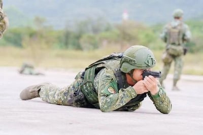 画面中一名身着全套迷彩作战服、头戴战术头盔的士兵呈卧姿趴在平整的硬质地面上，双手握持枪械，将枪械贴靠在脸颊旁，正专注地瞄准前方，似乎在进行射击训练。背景中还有其他身着同款军装的士兵，远处是模糊的绿植与开阔的户外环境，整体氛围严肃，展现出军事训练的严谨状态。