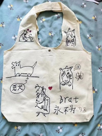 一张手绘帆布包的照片，包上有各种可爱的卡通图案，包括猫咪、狗狗和人物形象。包的左侧有一只被标注为'恶犬'的小狗，右侧有一只猫咪，猫咪的对话框里写着'汪'和'喵'，底部有一个卡通人物和'brat 永不为奴'的文字。整个设计风格可爱又带点幽默。