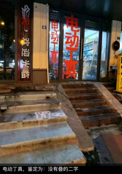 夜晚拍摄的一家商店门面，玻璃门上贴着醒目的红色"电动工具"字样，左侧有"今天，欢迎光临"的竖幅招牌。店门前有几级石阶，台阶上似乎有一些模糊的涂鸦。图片底部有一行文字："电动丁真，鉴定为：没有叠的二字"，玩了谐音梗，将"电动工具"的"动"和"丁真"联系起来制造笑点。