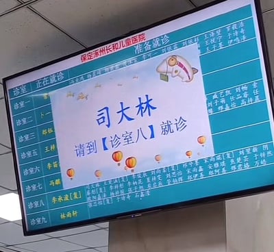 医院候诊区的电子显示屏，顶部显示'保定涿州长和儿童医院 准备就诊'，中间弹出提示框显示'司大林 请到【诊室八】就诊'，提示框内有卡通鱼形象和热气球装饰。屏幕左侧为各诊室信息，包括诊室一至九及对应医生姓名，右侧为患者名单列表。