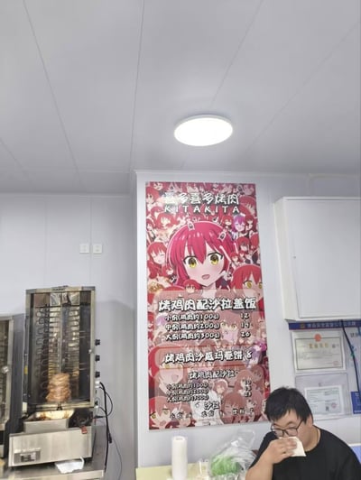 图片展示了一家餐厅的内部场景。左侧有一台不锈钢旋转烤鸡炉，里面正在烤制鸡肉。中间墙壁上挂着一幅大型动漫风格菜单海报，海报主体是一位红发黄眼的动漫女孩，周围环绕着多个相同角色的小头像，上方写有“壹多喜多样肉 KITAKITA”，下方列出了烤鸡肉配沙拉盖饭（小份12元、中份19元、大份26元）、烤鸡肉沙威玛卷饼（8元）、烤鸡肉配沙拉（小份10元、中份23元）等菜品信息，并标注沙拉、米饭、小菜可无限续。右侧有一位顾客坐在餐桌旁，正在用餐后用纸巾擦嘴，桌上放有餐具和食材。整体场景呈现出日常餐厅的温馨氛围，动漫元素的加入使菜单更具吸引力。