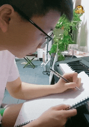 这是一段动态图，画面中戴眼镜的男生坐在钢琴旁，正低头专注地在笔记本上书写，右上角还添加了一个搞怪的小黄脸表情作为趣味点缀。背景里能看到绿植和一些室内物品，整体场景是日常的学习或创作时刻，没有谐音梗、双关或暗示内容。
