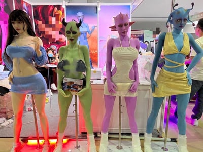 展会现场展示了四位身着时尚服装的外星风格模特，她们分别拥有蓝色、绿色、紫色和接近人类的肤色，搭配角饰、特殊妆容等科幻元素，造型前卫吸睛。背景可见展会海报和其他参观者，整体氛围充满奇幻色彩。模特们姿态各异，有的手持宣传册，有的单手叉腰，展现出自信专业的展示姿态。