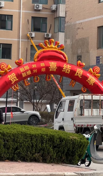 一张婚礼现场的照片，画面中央是一个红色的充气拱门，上面用金色字体写着“贺新郎刘超新娘刘超喜结良缘”以及英文“WEDDING”。拱门装饰有金色的皇冠造型和心形图案。背景是一栋多层砖楼，楼外有空调外机。拱门前方停着一辆白色卡车和一辆银色SUV，旁边有一位穿着深色衣服的人。整个场景呈现出喜庆的婚礼氛围，特别之处在于新郎新娘同名同姓，都叫刘超。