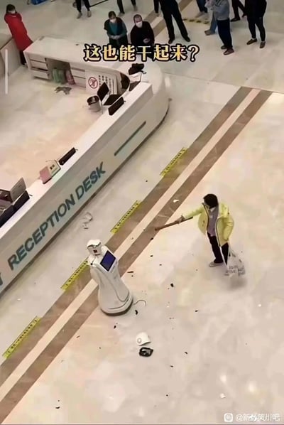 在标有“RECEPTION DESK”的服务大厅接待台前，一名身穿黄色外套的女子手持长棍，正在砸击一台白色服务机器人，机器人周围散落着被砸落的部件碎片，周围有多名围观人员。画面上方配文“这也能干起来？”，充满荒诞感，引发网友对冲突原因的好奇。