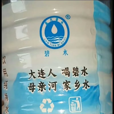 这是一张大连本土“蓝天碧水”品牌桶装水的标签实拍图，存在明显的视觉错位笑点：标签左侧竖排的文字本为“饮用纯净水”，但受字体、排版和反光效果影响，乍看很像“饮用纯海水”，搭配标签中间印的宣传语“大连人 喝碧水 母亲河 家乡水”，形成了调侃大连人喝海水的搞笑效果，这也是原帖标题《大连人有福了》的梗点来源。标签顶部还有蓝白色的品牌logo，印着水滴、水波纹图案以及“蓝天碧水”的对应拼音，底部还有可回收标识和垃圾投放提示标识。