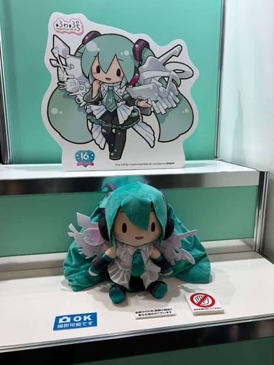 这是线下展示柜中的初音未来16周年限定周边，上层是带有翅膀装饰的Q版初音未来立牌，立牌标注了「ふわふち」系列、16周年纪念标识，以及画师和版权信息；下层是对应立牌形象的毛绒玩偶，玩偶还原了立牌的蓝绿色长发、耳机、翅膀与服饰设计。展示台配有日文标识：蓝色标识表示可以拍照，禁止触摸标识提示不要触碰，还有提示牌说明该商品处于监修中，可能与最终商品有差异。