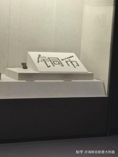 这是博物馆玻璃展柜内的创意展品布置，工作人员用多枚古铜币在展示台的白色斜面展板上拼出「铜币」两个汉字，直观点明展品主题；旁边的展示台还陈列着一件小型骨制文物，整体展陈空间简洁规整，用展品本身做创意设计，形式新颖有趣。
