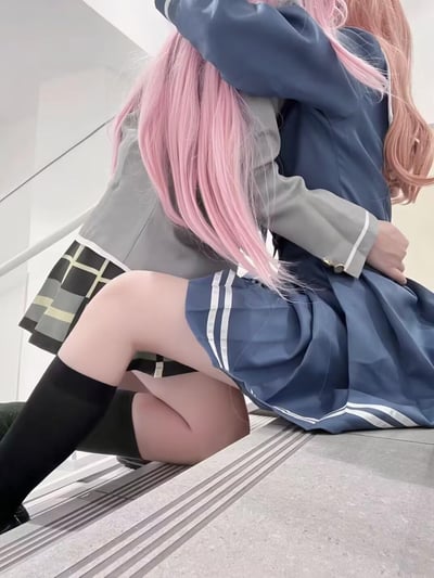 图片展示了两个穿着日式校服的女孩在楼梯上拥抱的场景。左边的女孩有着粉色长发，穿着灰色西装外套和黑色格子裙，搭配黑色过膝袜。右边的女孩有着浅粉色长发，穿着蓝色西装外套和蓝色百褶裙，裙子上有白色条纹装饰。两人背对着镜头，坐在楼梯上相互拥抱，姿态亲密，展现出温馨的氛围。背景是简洁的白色建筑结构，突出人物主体。