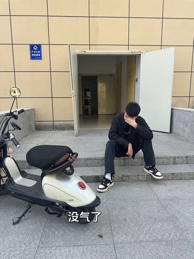 这张竖版实拍图展示了建筑物入口的户外场景，墙面贴有“严禁占用 消防通道”的蓝色标识，一名穿黑衣黑裤的年轻男子坐在入口台阶上，单手捂脸情绪低落，他面前停着一辆轮胎没气的电动自行车，图片下方配文“没气了”。内容利用文字双关制造笑点，“没气了”既指代电动车轮胎漏气，也很容易让第一眼看到的人误以为是旁边的男子出意外失去生命体征，调侃这种歧义会吓到负责学生安全的高校辅导员。