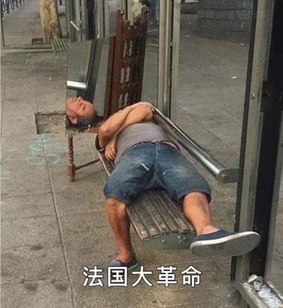 图片中一名男子以放松的姿势横躺在公交站旁的长椅上，双臂交叉抱于胸前，双腿伸直搭在长椅边缘，头部靠在椅背，似乎在沉睡。长椅位于人行道旁，背景可见公交站的玻璃挡板和街道地面。图片底部有白色文字“法国大革命”，形成一种荒诞的幽默对比——将历史上激烈的革命事件与男子此刻“躺平”般的悠闲状态并置，制造出无厘头的笑点。