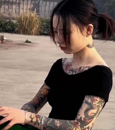 画面是一位身穿黑色短袖上衣的年轻女性，坐在户外简单地背景下，展示出满臂以及脖子、肩膀上复杂且色彩丰富的纹身。整体气质非常酷，有视觉冲击力，纹身包含许多花纹与图案，表现出叛逆个性、艺术感或自我表达。整体画面干净，女孩侧坐，显得安静且自信，给人一种自由、不受拘束的感觉。