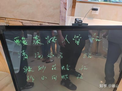 这是一张在餐厅内实拍的手写黑板菜单照片，深色玻璃黑板上用绿色荧光笔手写了四道家常菜及对应价格：<br>1. 酸菜炖大骨 6元<br>2. 清汤丸子 8元<br>3. 辣炒花蛤 4元<br>4. 炸小河虾 6元<br>黑板表面能看到餐厅内顾客的人影倒影，背景是餐厅的柜台与石质墙面，整体呈现出家常小馆的接地气风格，菜品定价十分实惠。图中无谐音梗、双关或暗示类内容。