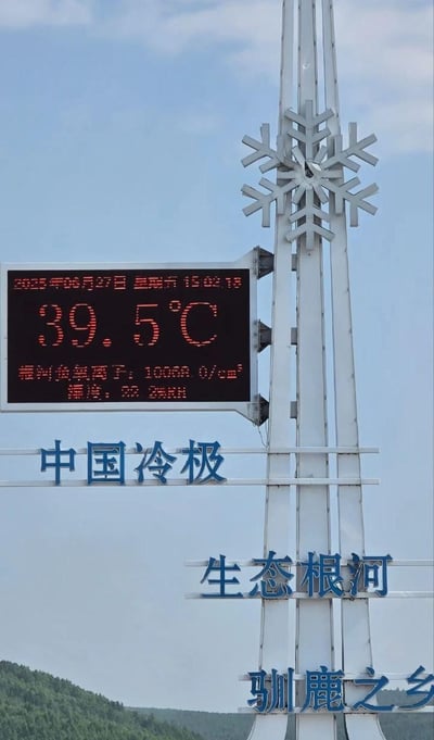图片展示了一个高耸的金属结构标牌，顶部为雪花造型装饰，下方电子屏显示2025年06月27日15:02:18的实时数据：气温39.5℃、空气负氧离子10000个/cm³、风力2.0m/s。标牌主体有蓝色文字“中国冷极”“生态根河”“驯鹿之乡”，背景为晴朗天空和远处的绿色山林。作为以“冷极”为标志的地区出现近40℃高温，形成鲜明的视觉与概念反差。