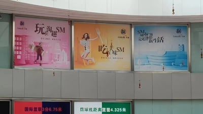 图片展示了某商业建筑外墙上的三块并排广告牌，整体为横向构图。左侧粉色广告牌标题为“玩淘住趣 SM”，画面中有一位提着购物袋奔跑的人物，背景有建筑剪影；中间橙色广告牌标题为“吃定美味 SM”，一位女性模特手持刀叉跳跃，姿态活泼；右侧蓝色广告牌标题为“SM城市广场 定义新生活”，背景是抽象的建筑与生活场景。广告牌下方另有两块小型标牌，分别标注“国际篮联3分6.75米”和“罚球线距离篮筐4.325米”。整体风格充满商业宣传气息，色彩鲜明，人物形象富有活力。