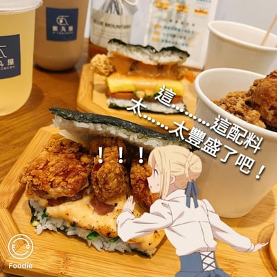 图片展示了一张美食照片，前景是两个放在木制托盘上的米饭三明治，里面夹着大量炸鸡块和其他配料。一个金发动漫女性角色被P在图片下方，表情惊讶地伸手触碰食物。图片上有文字标注："這配料...太...太豐盛了吧！"和"!!!"，表达对食物分量的惊叹。背景中可以看到饮料和其他食品包装。