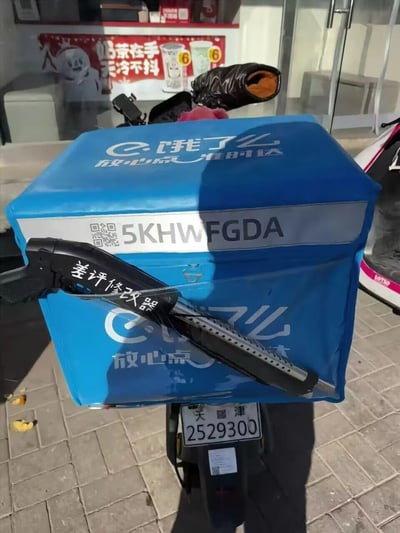这是一张户外实拍的静态照片，主体是饿了么品牌的蓝色外卖保温箱，箱体上印有“饿了么 放心点”的品牌标识，中部银灰色条带区域印有编号“5KHWF GDA”和对应二维码，顶部也印有“饿了么 放心点 准时达”的字样。外卖箱上用绑带固定了一把黑色玩具枪，枪身印有白色的“差评修改器”字样，是外卖员为调侃行业差评压力制作的趣味道具。背景可见蜜雪冰城门店，玻璃上有“奶茶在手 天冷不抖”的宣传语，外卖箱下方是悬挂天津牌照（2529300）的电动车，拍摄场景为户外砖地路面，整体氛围轻松搞笑，用夸张道具调侃外卖行业的职场压力。