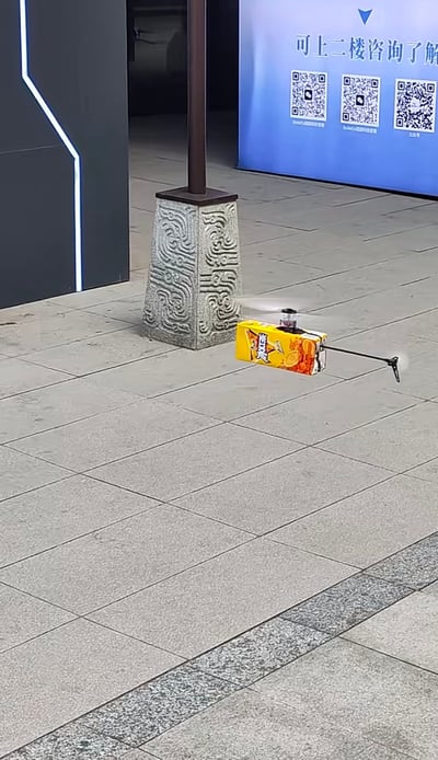 这是一张户外实拍静态照片，画面主体是一架由冰红茶黄色外包装盒手工改装而成的小型无人机，正处于飞行状态，旋翼因转动呈现模糊效果。拍摄场景为铺有灰色方形地砖的户外公共区域，背景可见带有中式传统云纹类纹样雕刻的石质灯柱底座，还有一块蓝色宣传展板，整体呈现出创意十足的手工改装趣味效果。